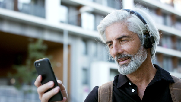 Portrait of happy mature man with headphones and smartphone outdoors in city, listening to music.