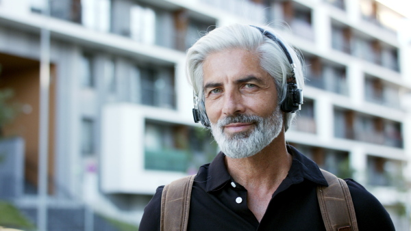 Portrait of mature man with headphones outdoors in city, using smartphone and looking at camera.