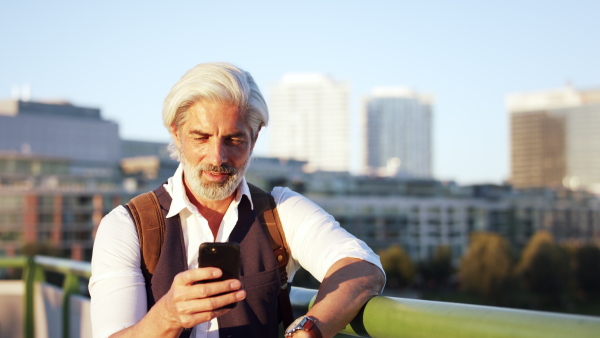 Front view portrait of mature man standing outdoors in city, using smartphone.