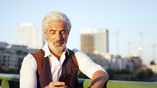 Front view portrait of mature man standing outdoors in city, using smartphone.