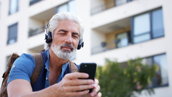 Portrait of mature man with smartphone and headphones sitting outdoors in city, listening to music.