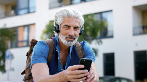 Close-up portrait of mature man with headphones sitting outdoors in city, using smartphone.