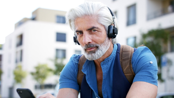 Portrait of mature man with smartphone and headphones sitting outdoors in city, looking at camera.