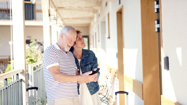 Happy senior couple outside apartment on holiday, using smartphone.