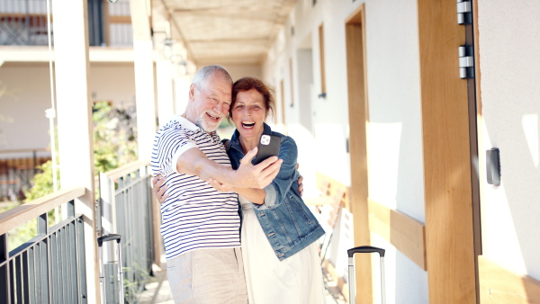 Cheerful senior couple outside apartment on holiday, taking selfie with smartphone.