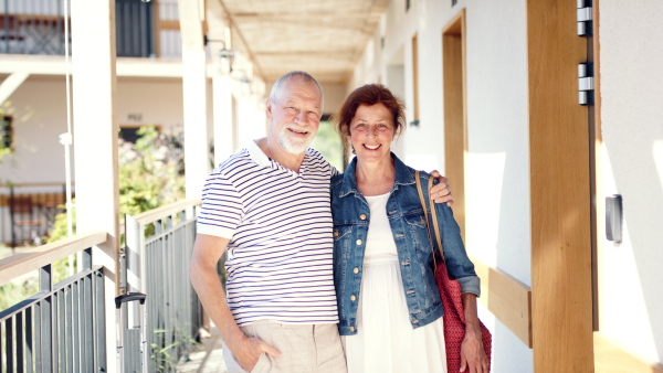 Portrait of senior couple with luggage outside apartment on holiday, looking at camera.