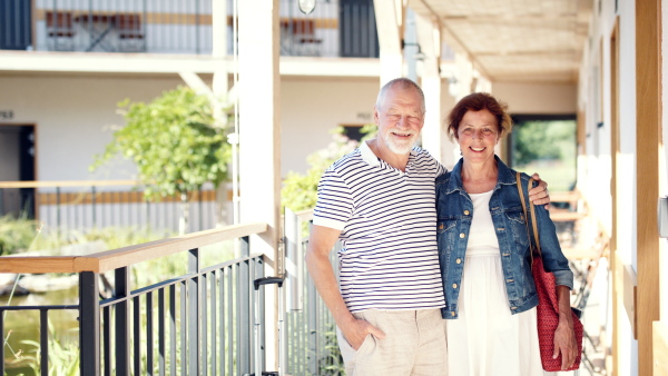 Portrait of senior couple with luggage outside apartment on holiday, looking at camera.