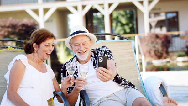 Happy senior couple with wine on sunbeds outdoors on holiday, taking selfie.