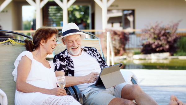 Cheerful senior couple with wine on sunbeds outdoors on holiday, using tablet.