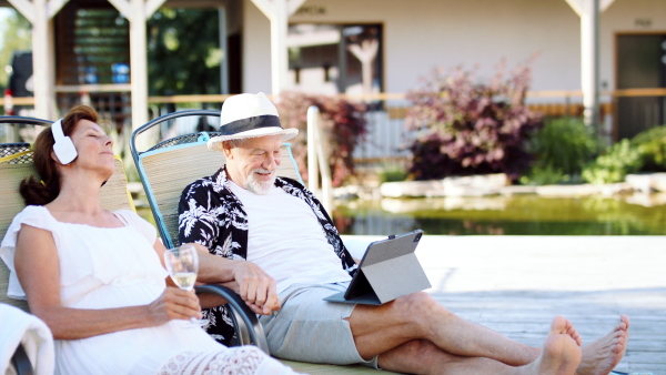 Front view of senior couple with tablet and headphones outdoors on holiday, relaxing.