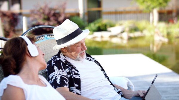 Front view of senior couple with tablet and headphones outdoors on holiday, relaxing.