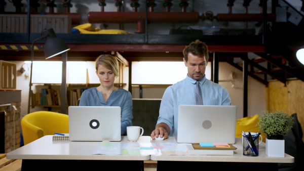 Portrait of man and woman sitting indoors at desk in office, working.