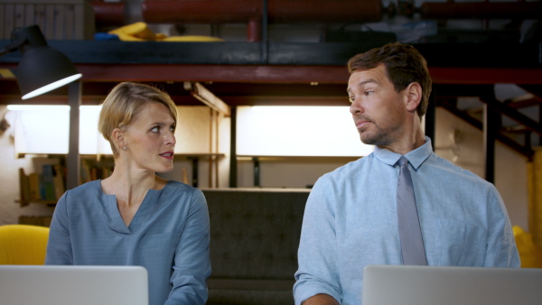 Portrait of man and woman with laptop sitting indoors at desk in office, working.