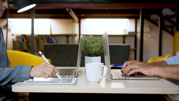 Unrecognizable man and woman sitting indoors at desk in office, working with laptop and tablet.