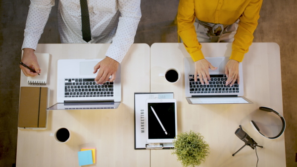 Top view of unrecognizable man and woman sitting indoors at desk in office, working with laptops.