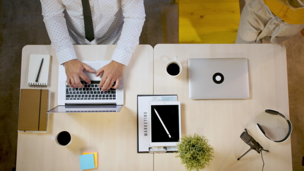 Top view of unrecognizable man and woman sitting indoors at desk in office, working with laptops.