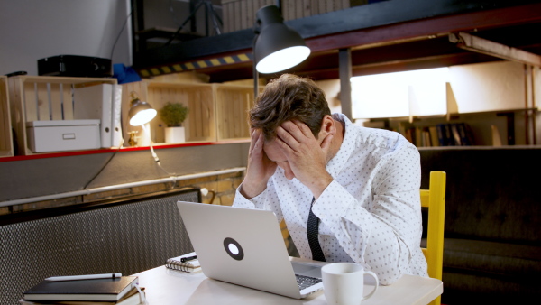 Sad and frustrated businessman with laptop sitting indoors at desk in home office, working.