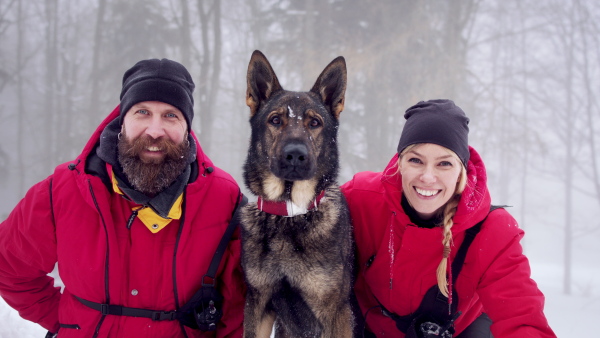 Mountain rescue service with dog on operation outdoors in winter in forest, celebrating and looking at camera.