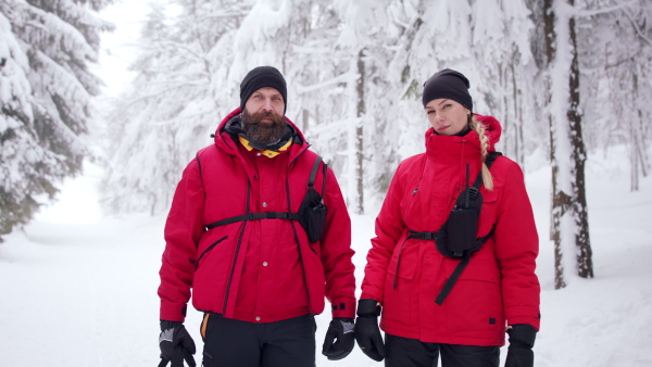 Man and woman paramedics from mountain rescue service outdoors in winter in forest, looking at camera.