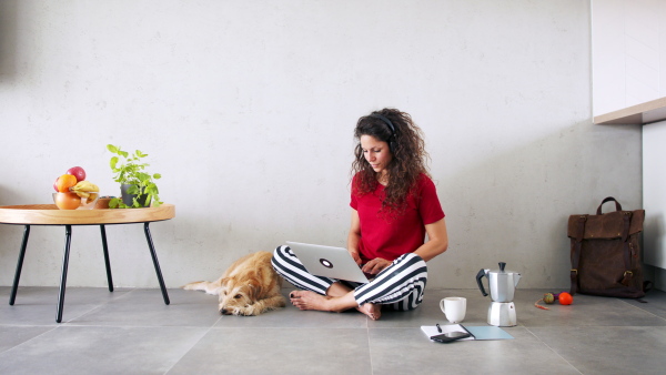 Portrait of woman with pet dog and headphones indoors at home, using laptop.