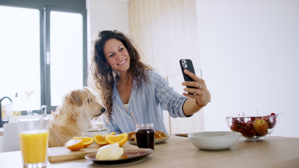 Portrait of woman with pet dog taking selfie indoors at home during breakfast.