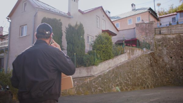 A rear view of delivery man courier delivering parcel box in town using smartphone.