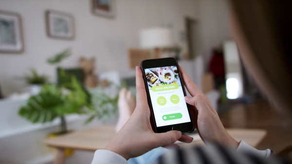 Rear view of young woman with smartphone ordering food at home, coronavirus and food delivery concept.