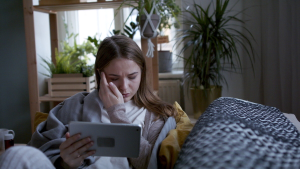 Worried young woman wrapped in blanket using tablet at home, coronavirus concept.