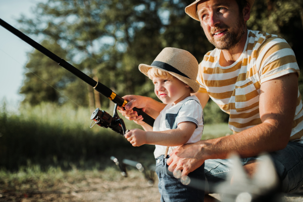 A mature father with a small toddler son outdoors fishing by a river or a lake.