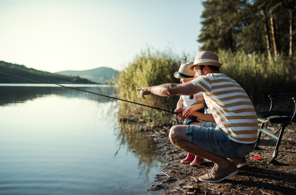 A mature father with a small toddler son outdoors fishing by a river or a lake.