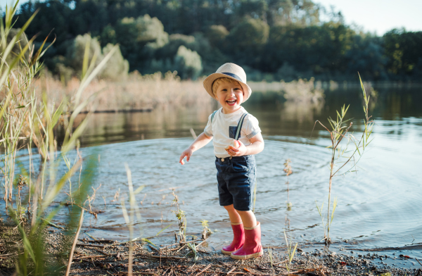 A cheerful small toddler boy standing by a lake, eating an apple.