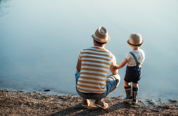 A rear view of father with a small toddler son spending time outdoors by a lake. Copy space.