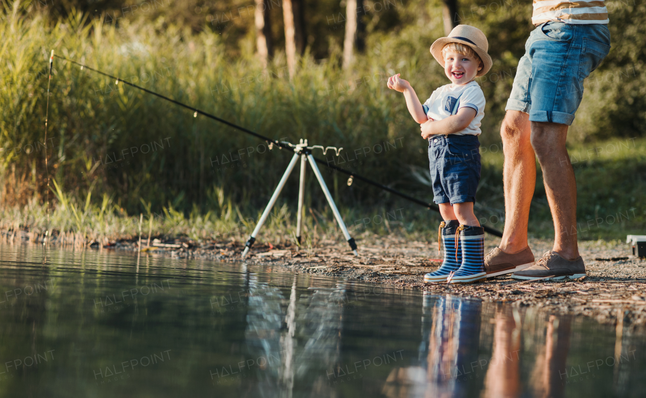 An unrecognizable father with a small toddler son outdoors fishing by a river or a lake.