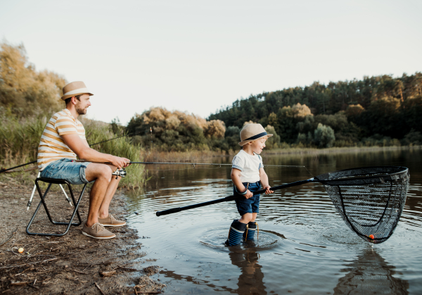 A mature father with a small toddler son outdoors fishing by a river or a lake.