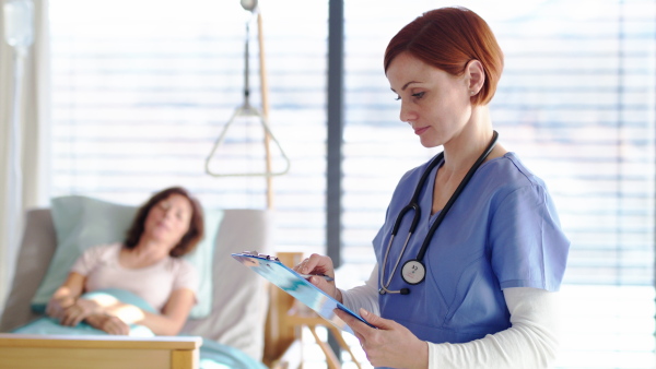 Friendly woman doctor or nurse examinig patient in bed in hospital, making notes.