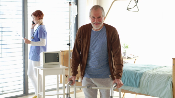 A senior man patient with walking frame and nurse in hospital room.