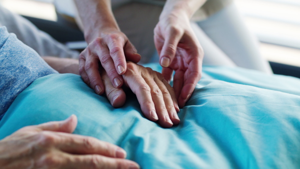 Midsection of unrecognizable friendly woman doctor sitting by patient in bed in hospital, holding hands.