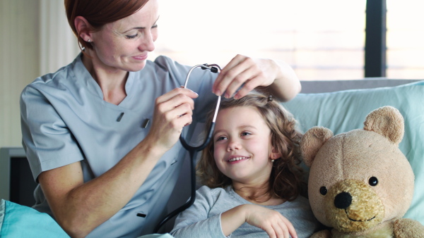 Friendly woman doctor with stethoscope examining small girl in bed in hospital.