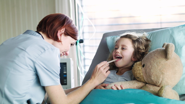 Friendly woman doctor with dongue depressor examining small girl in bed in hospital.