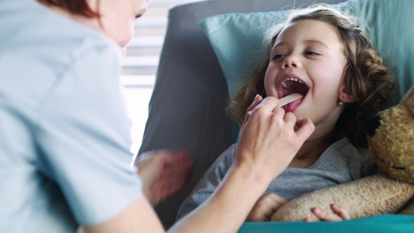 Female doctor with tongue depressor examining small girl in bed in hospital.