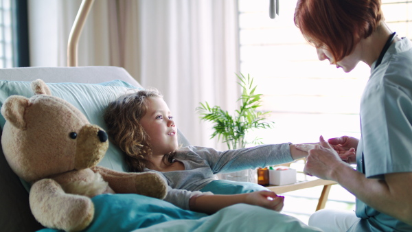 Friendly female doctor examining small girl in bed in hospital, talking.