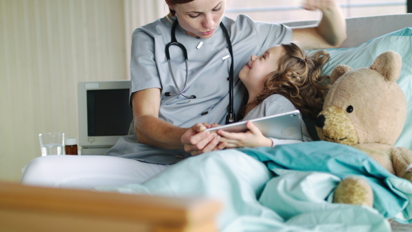 Friendly female doctor and small girl in bed in hospital, using tablet. and talking.