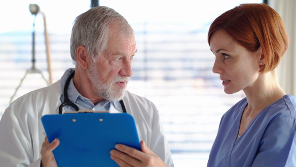 Senior male doctor with clipboard standing in hospital room, talking to a nurse.