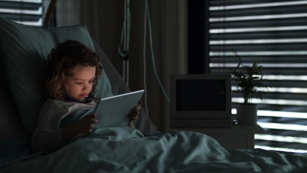 Bored small girl lying in bed in hospital, using tablet.