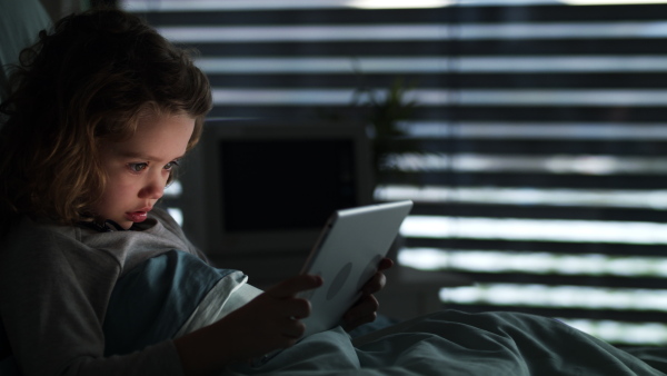 Bored small girl lying in bed in hospital, using tablet.