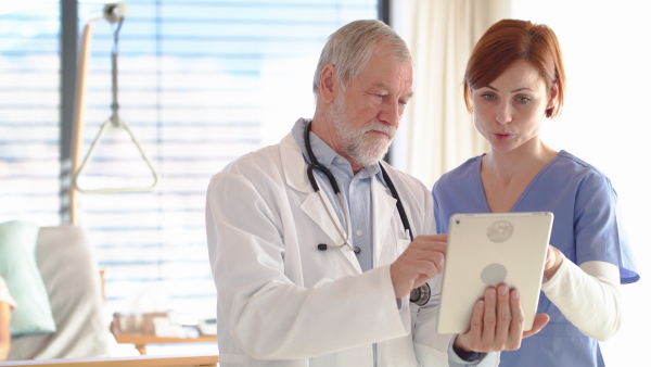 Senior male doctor with tablet standing in hospital room, talking to a nurse.