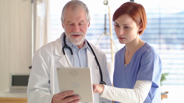 Senior male doctor with tablet standing in hospital room, talking to a nurse.