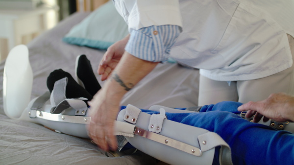 Unrecognizable healthcare worker and man paralysed senior patient in hospital, applying orthosis.