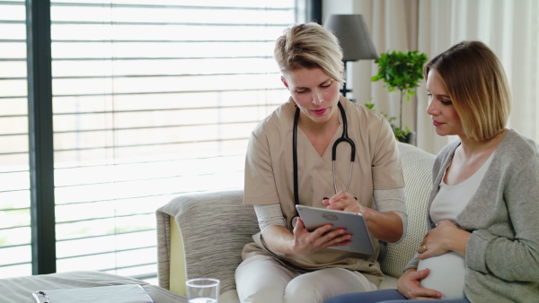 Healthcare worker with tablet and pregnant woman on sofa at home, talking.