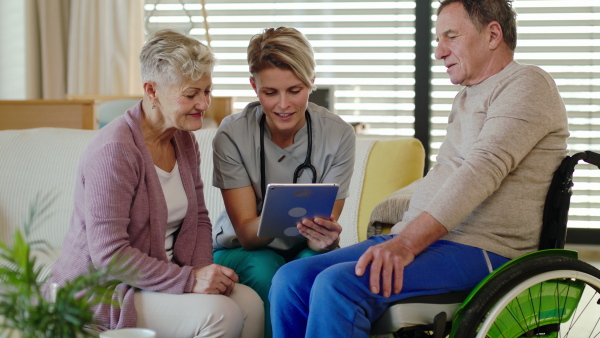 Healthcare worker with tablet visiting senior patient in wheelchair at home, talking.